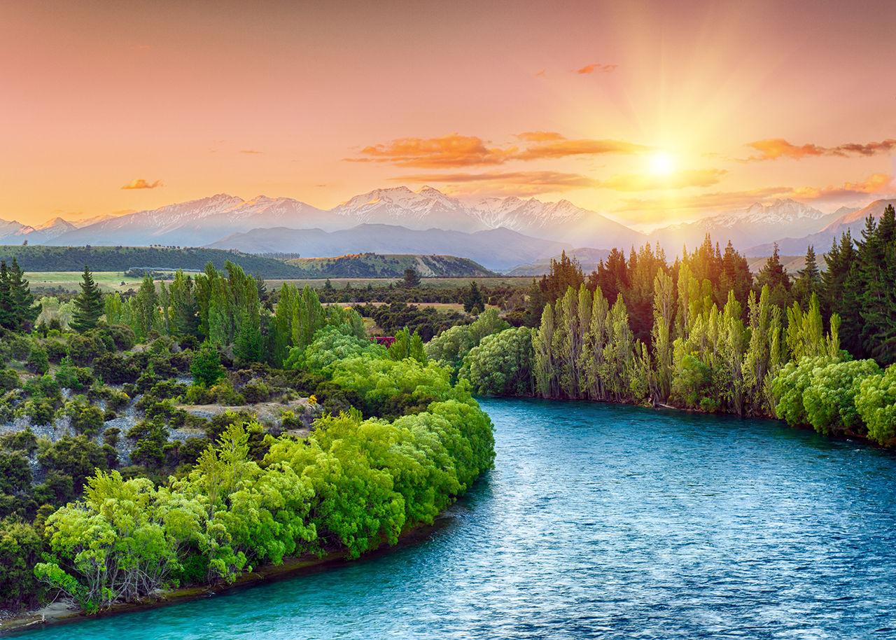 River running through a forested scene with mountains and a sunset in the background
